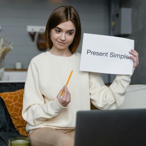Lektorka Výuky jazyků online vede online výuku angličtiny a ukazuje na notebooku papír s tématem Present Simple.